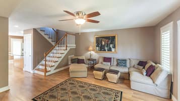 A spacious living room with a wooden staircase, a ceiling fan, a large framed wine bottle artwork, and a patterned rug.