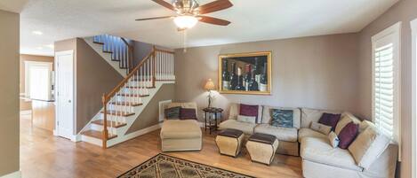 A spacious living room with a wooden staircase, a ceiling fan, a large framed wine bottle artwork, and a patterned rug.