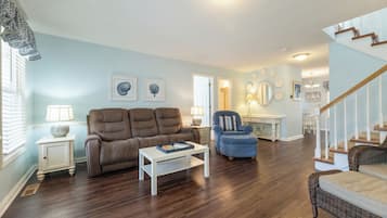 Ein Wohnzimmer mit braunem Sofa, blauem Sessel und weißem Couchtisch. Eine Treppe befindet sich rechts, und eine Tür führt in einen weiteren Raum.