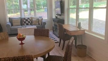 Ein Essbereich mit Holztisch und Korbstühlen, einer Couch mit Kissen, einem Fernseher und großen Fenstern mit Blick auf Grünflächen.