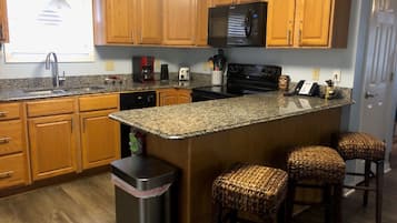 A kitchen with wooden cabinets, a granite countertop, a microwave, a stove, a coffee maker, and a trash can. There are three wicker barstools.