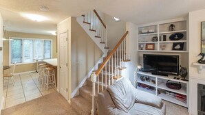 A living room with a beige couch, a TV, and a bookshelf. There is a staircase with wooden railings leading to another room. - Three Bedroom Hurst Haven Condo (Grand Rivers)