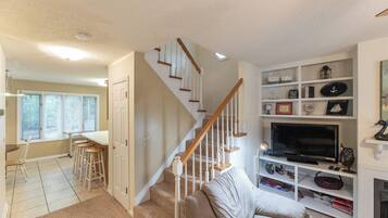 A living room with a beige couch, a TV, and a bookshelf. There is a staircase with wooden railings leading to another room.