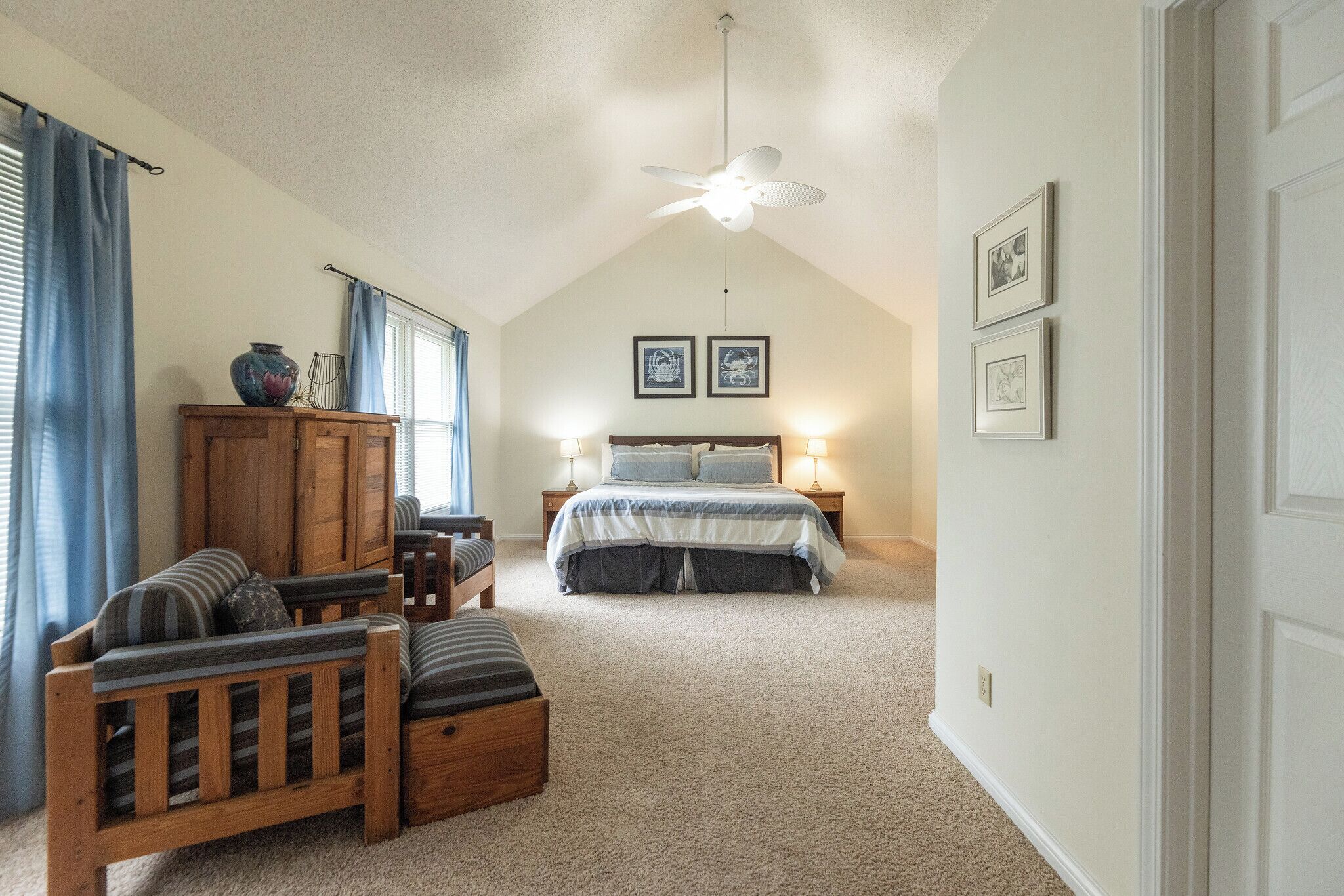 A bedroom with a sloped ceiling, a ceiling fan, a bed with a blue and white bedspread, two bedside tables with lamps, a wooden cabinet, a wooden bench, and a window with blue curtains.