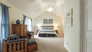 A bedroom with a sloped ceiling, a ceiling fan, a bed with a blue and white bedspread, two bedside tables with lamps, a wooden cabinet, a wooden bench, and a window with blue curtains. - Three Bedroom Hurst Haven Condo (Grand Rivers)