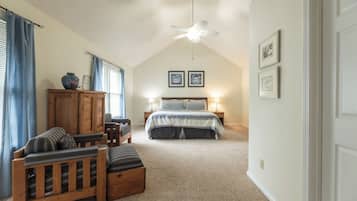 A bedroom with a sloped ceiling, a ceiling fan, a bed with a blue and white bedspread, two bedside tables with lamps, a wooden cabinet, a wooden bench, and a window with blue curtains.
