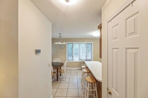 Une cuisine avec une table à manger et des tabourets de bar, une horloge au mur et une fenêtre avec des stores.