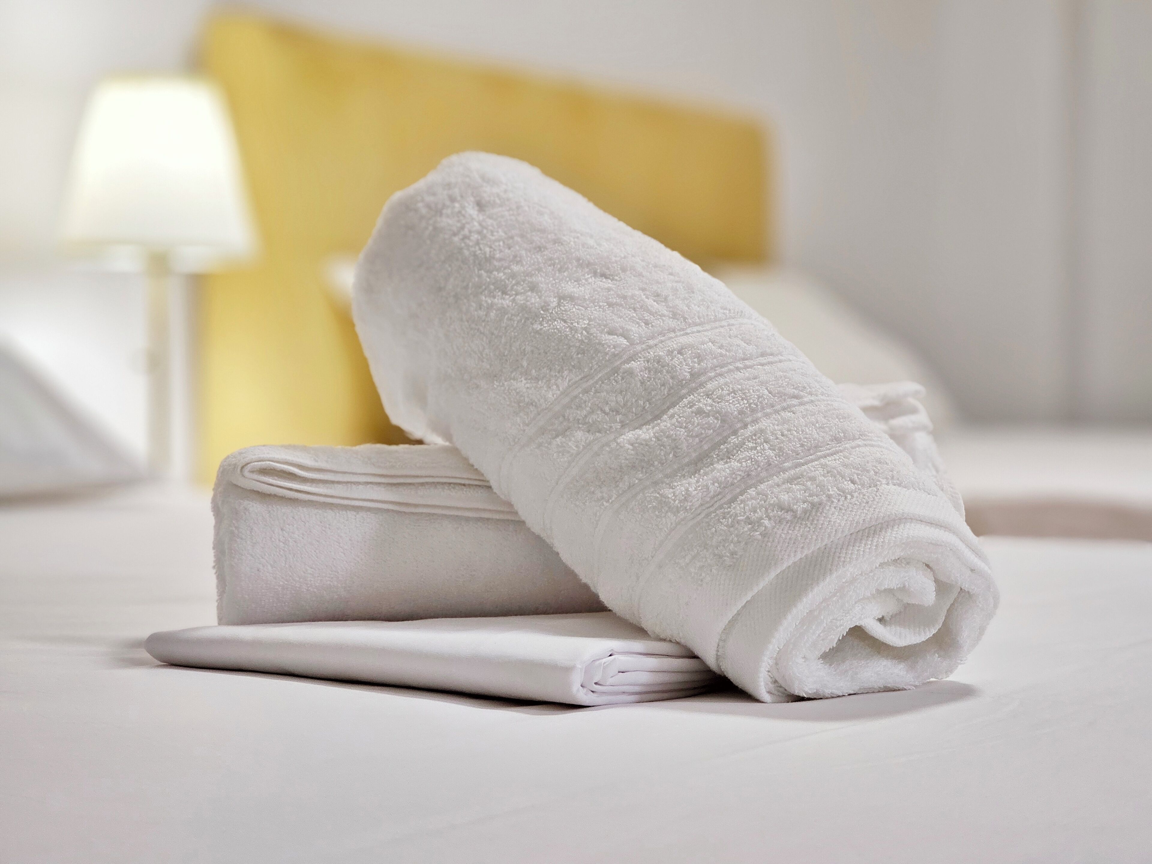 A neatly folded white towel on a bed with a white pillow and a white headboard.