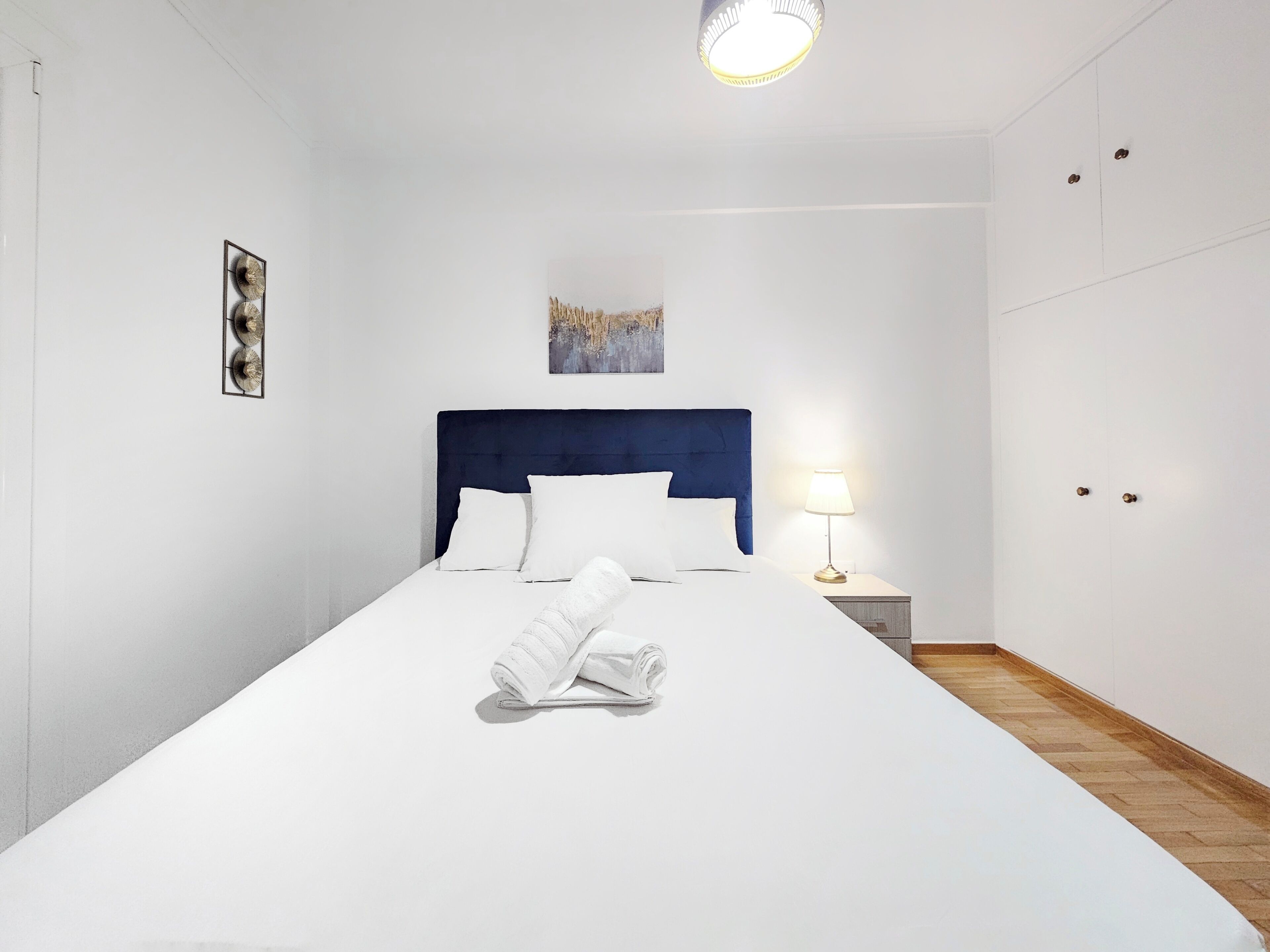 Een net opgemaakt bed met witte lakens, een blauw hoofdeinde en een bedlampje.