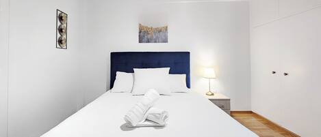 Een net opgemaakt bed met witte lakens, een blauw hoofdeinde en een bedlampje.