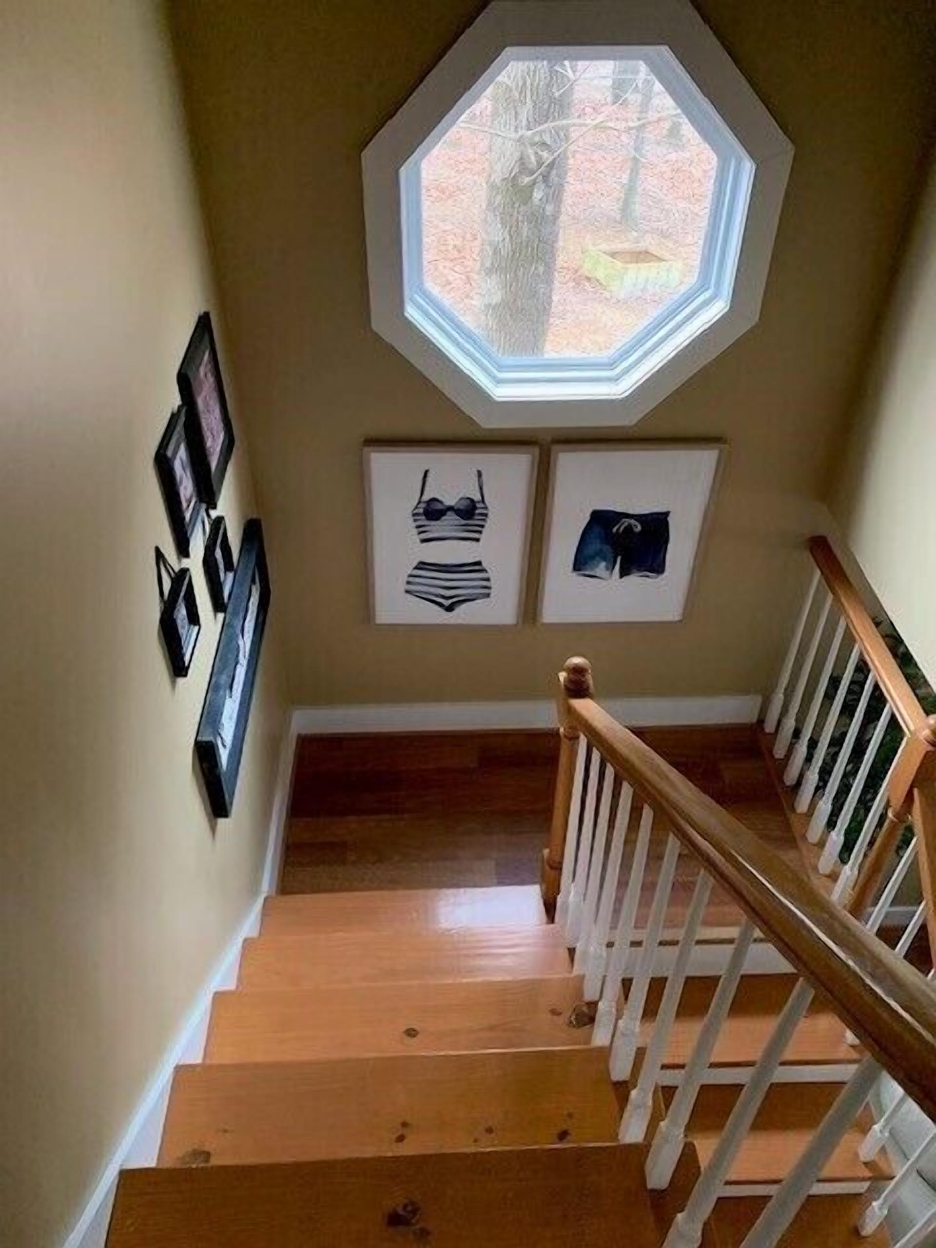 A wooden staircase with a white railing, a large octagonal window, and framed pictures on the wall.
