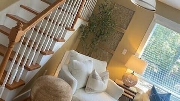 Een ruime woonkamer met een houten trap, een gezellige zithoek met een bank en voetenbankje, een bijzettafeltje met een lamp, en een groot raam dat voor natuurlijk licht zorgt.