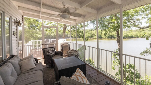 A covered porch with a view of a lake, furnished with a sofa, chairs, and a table.
