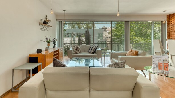 Una sala de estar moderna con un sofá, sillones y una mesa de centro de vidrio. A la izquierda, hay un aparador de madera y una planta. La habitación tiene ventanas amplias con vistas a edificios y árboles.