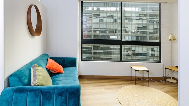 A modern living room with a teal sofa, a round mirror, a large window with blinds, a small table, and a lamp. - Perfect 1 BR in Centro Historico (Bogotá)