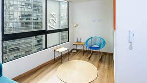 A modern room with a large window, a small table, a blue chair, a floor lamp, and a woven rug. - Perfect 1 BR in Centro Historico (Bogotá)