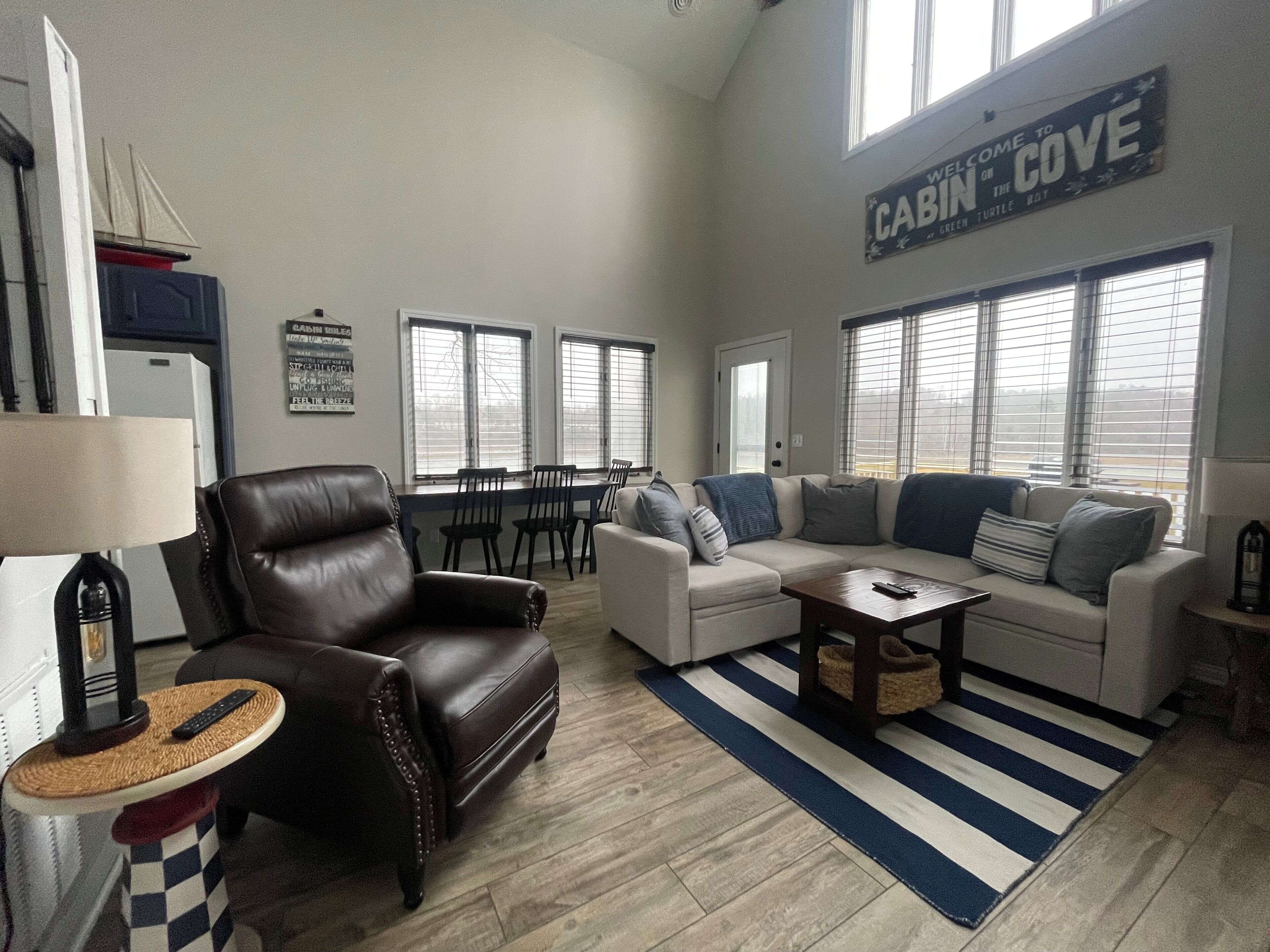 A spacious living room with a leather armchair, a white sofa, and a striped area rug. There are multiple windows providing natural light, and a sign above the sofa reads "Welcome to Cabin Cove."