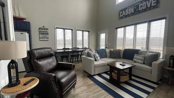 A spacious living room with a leather armchair, a white sofa, and a striped area rug. There are multiple windows providing natural light, and a sign above the sofa reads "Welcome to Cabin Cove."