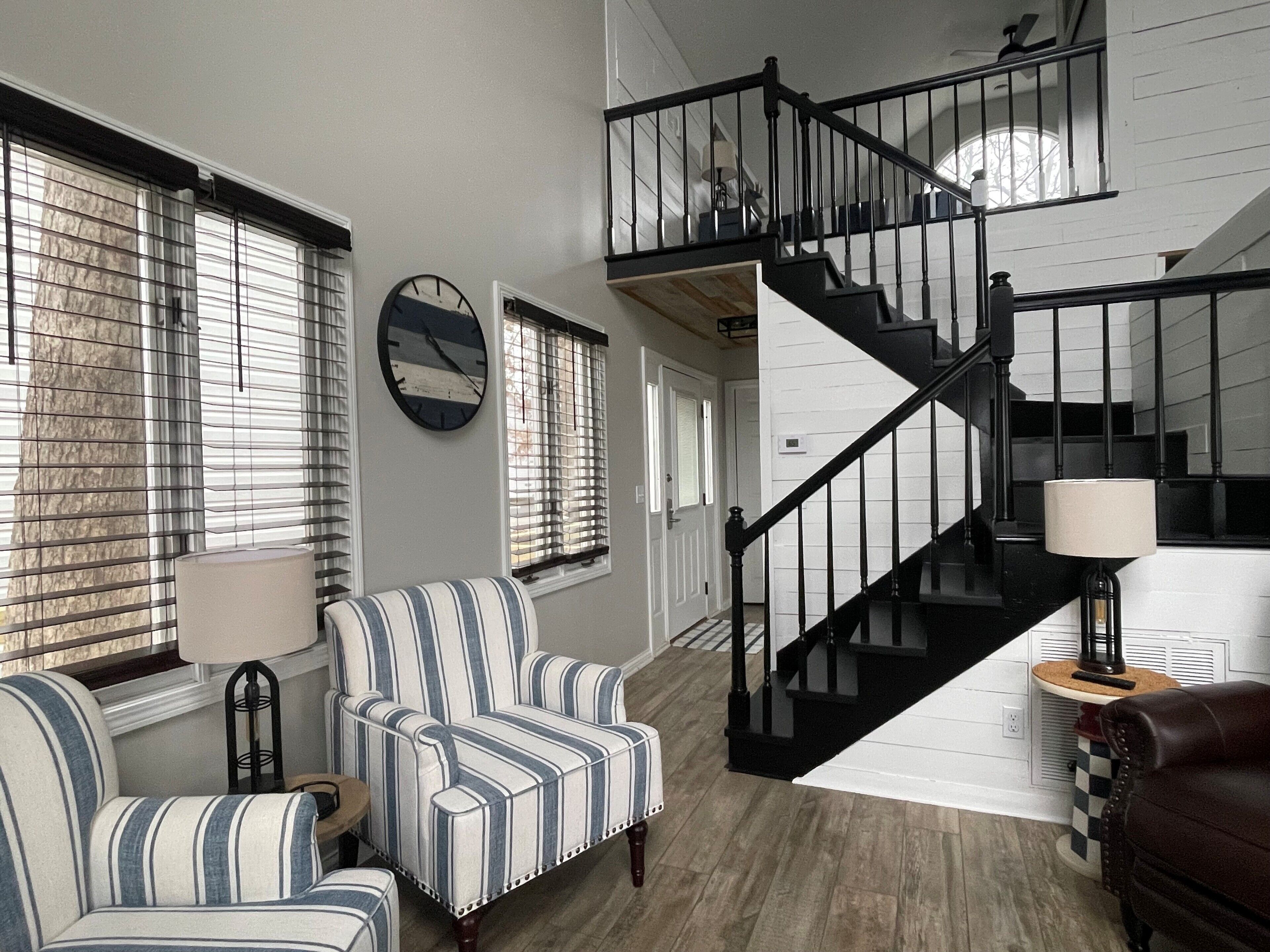 A spacious living room with a striped armchair, a staircase with black railings, and a large clock on the wall.