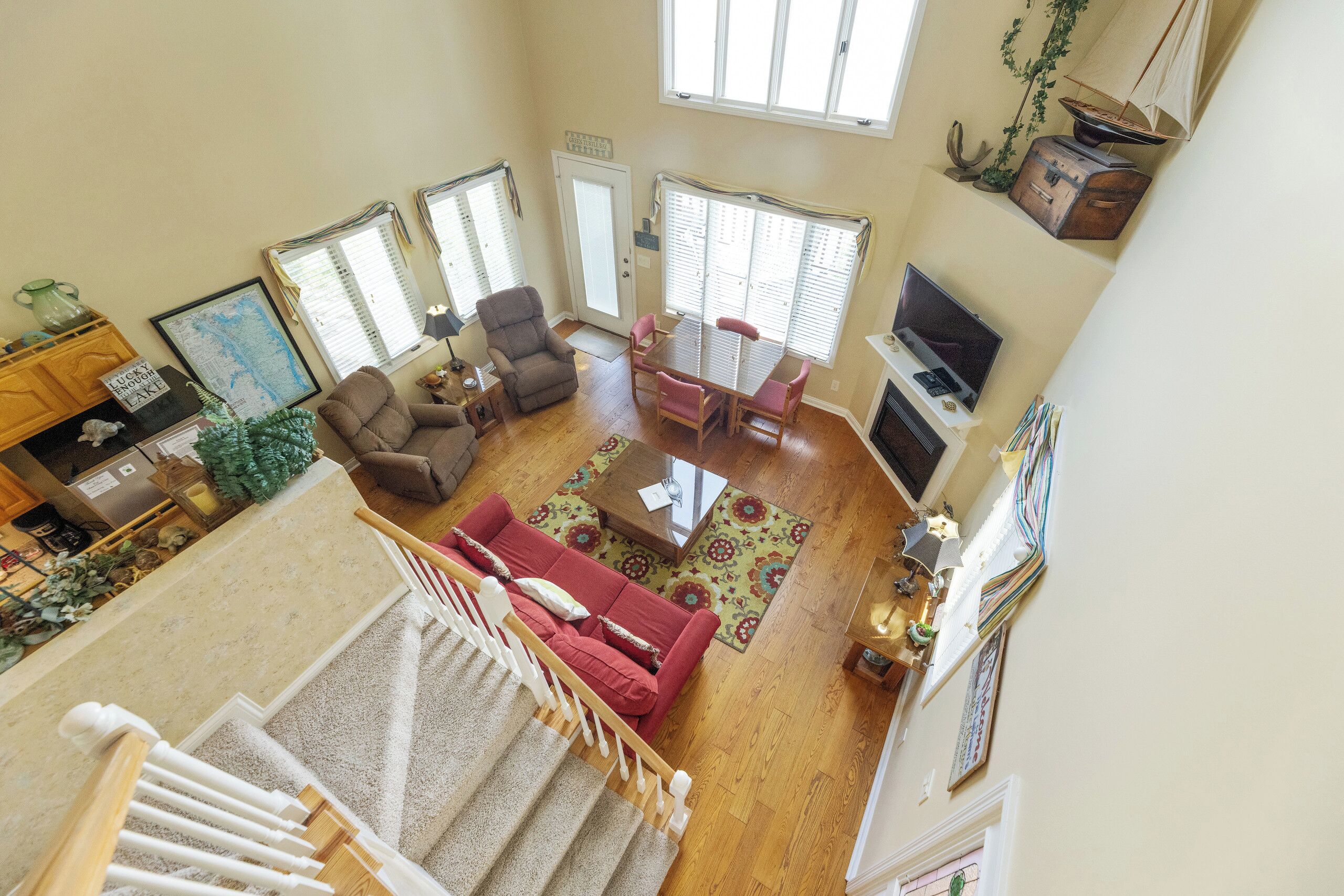 A spacious living area with a red sofa, a wooden coffee table, and a TV. There are two windows with blinds, a staircase, and a door leading to another room.