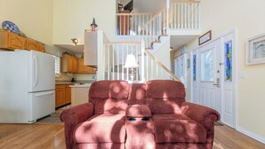 A spacious living room with a large, plush red sofa in the foreground, a kitchen with wooden cabinets and a white refrigerator, and a staircase leading to an upper level.