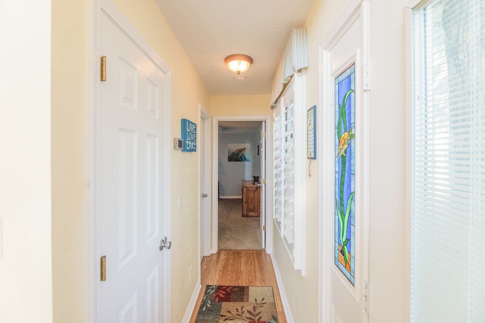 A hallway with white doors, a wooden floor, and a rug leading to a room with a painting and a window with blinds.