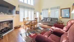 A spacious living room with a fireplace, a television, a dining area with a round table and chairs, a sofa, and a large window with blinds.