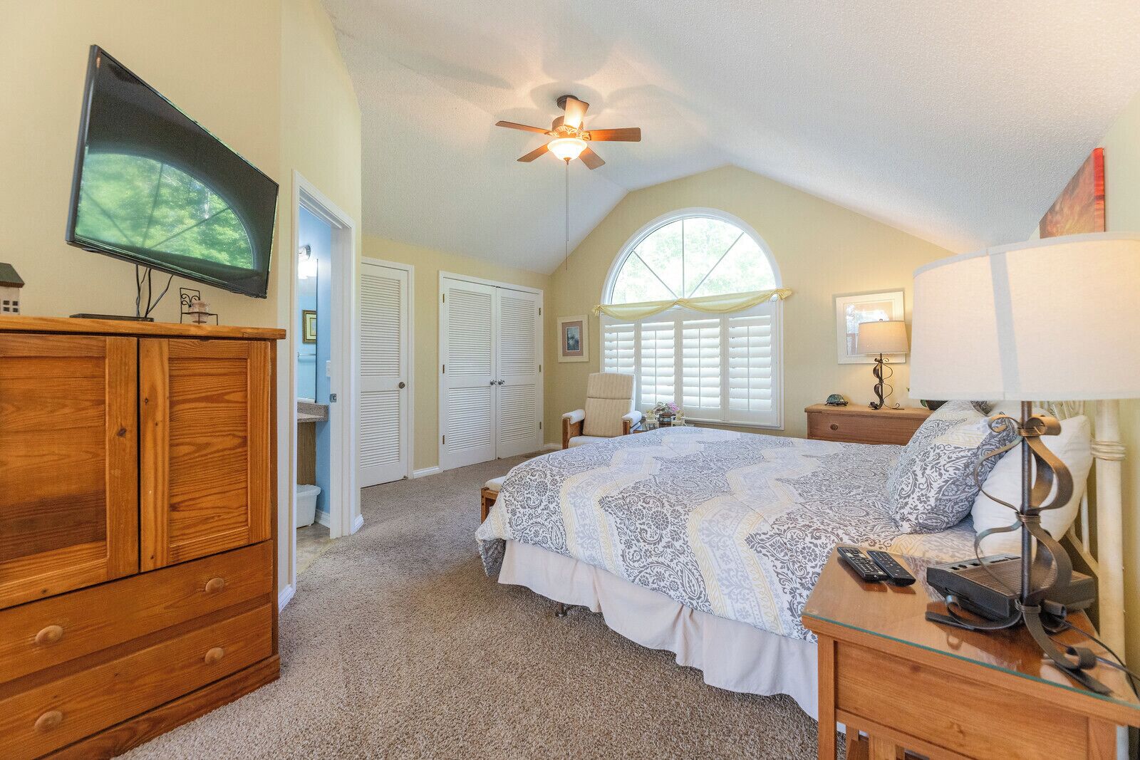 A bedroom with a large bed, a ceiling fan, a television, and a window with shutters.