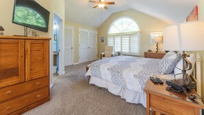 A bedroom with a large bed, a ceiling fan, a television, and a window with shutters.