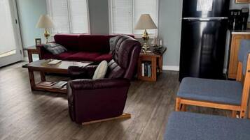 A living room with a sofa, chairs, a dining table, and a kitchen area with a refrigerator.