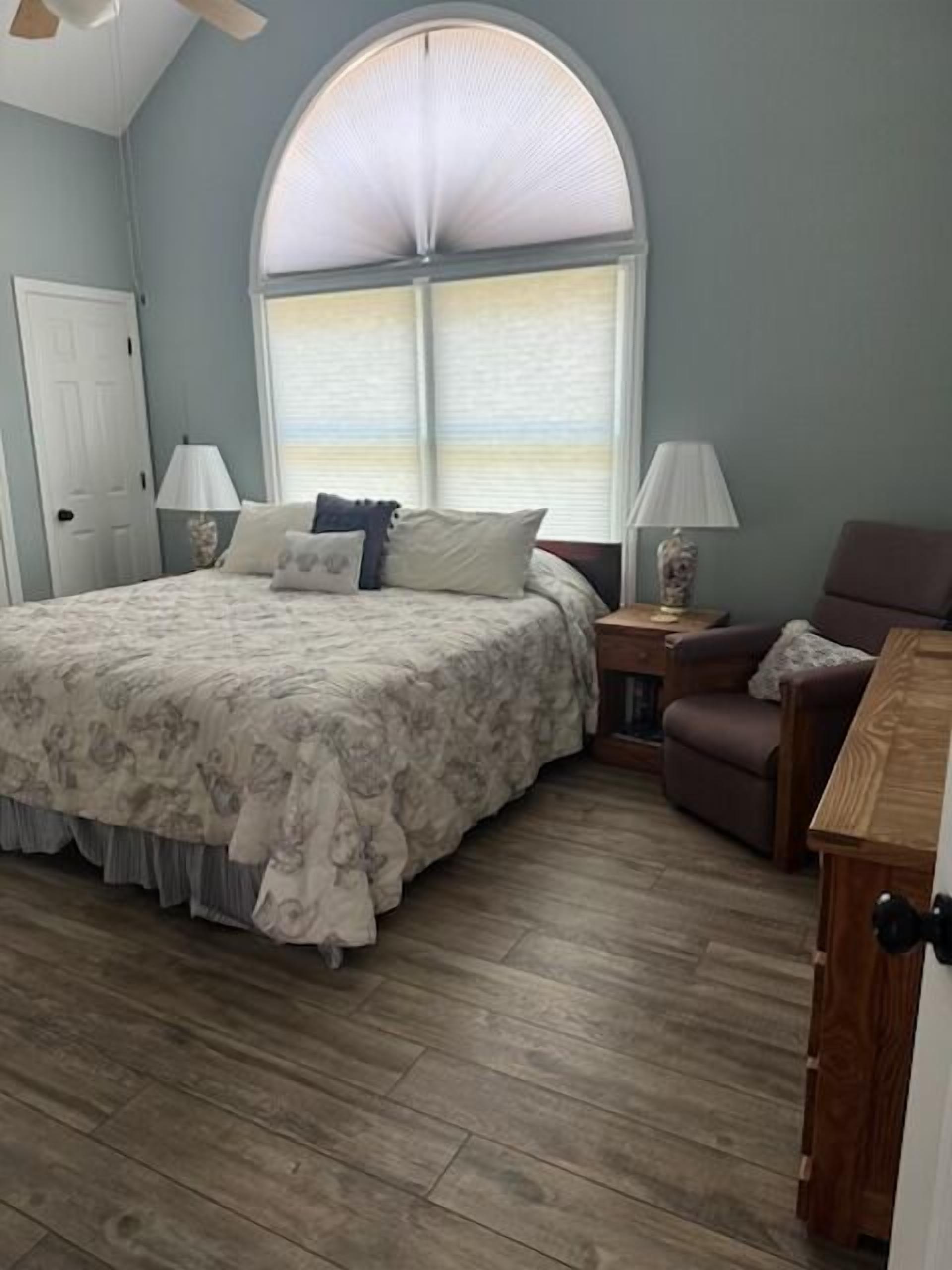 A bedroom with a large bed, a wooden floor, a window with blinds, a nightstand, and a chair.