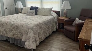 Una camera da letto con un letto grande, pavimento in legno, una finestra con persiane, un comodino e una sedia.