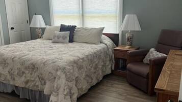 A bedroom with a large bed, a wooden floor, a window with blinds, a nightstand, and a chair.