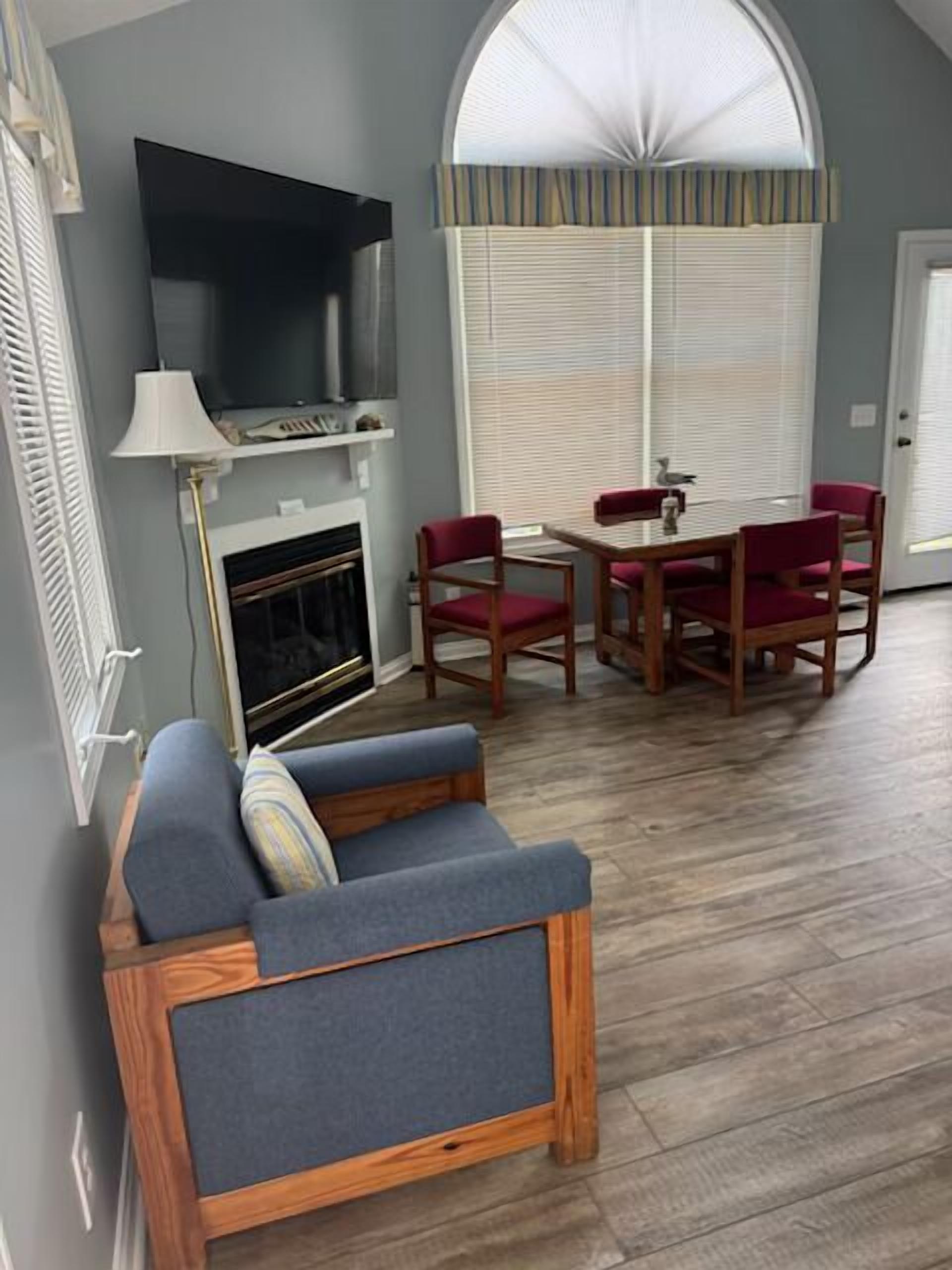 A room with a fireplace, a television, a dining table with chairs, and a large window with blinds.