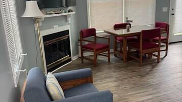 A room with a fireplace, a television, a dining table with chairs, and a large window with blinds.