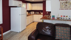 A compact kitchen with white cabinets, a refrigerator, microwave, and a stove. There's a brown leather couch with a patterned pillow. - One Bedroom Bayside Condo (Grand Rivers)