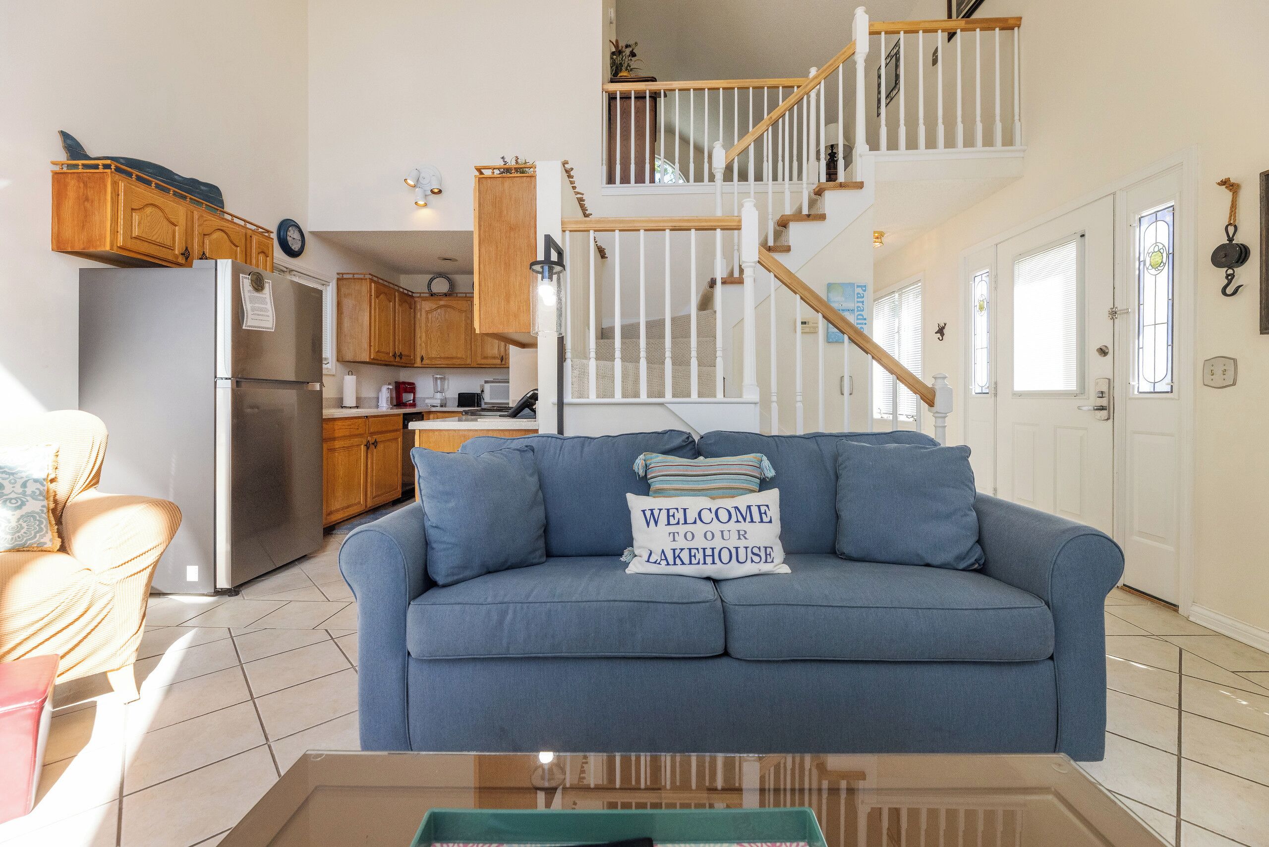 A living room with a blue sofa, a kitchen in the background, and a staircase leading to an upper level.