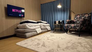 A room with a bed, a chair, a TV displaying 'NETFLIX', a lamp, and a small table with items on it.