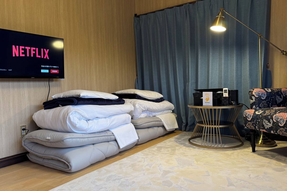Ein Hotelzimmer mit einem Bett, einem Fernseher, der 'NETFLIX' anzeigt, einem Nachttisch mit Lampe und einem gemusterten Sessel.