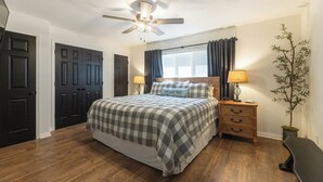 A bedroom with a bed, a ceiling fan, a nightstand with a lamp, a potted plant, and a window with curtains. - Two Bedroom Bayside Condo (Grand Rivers)