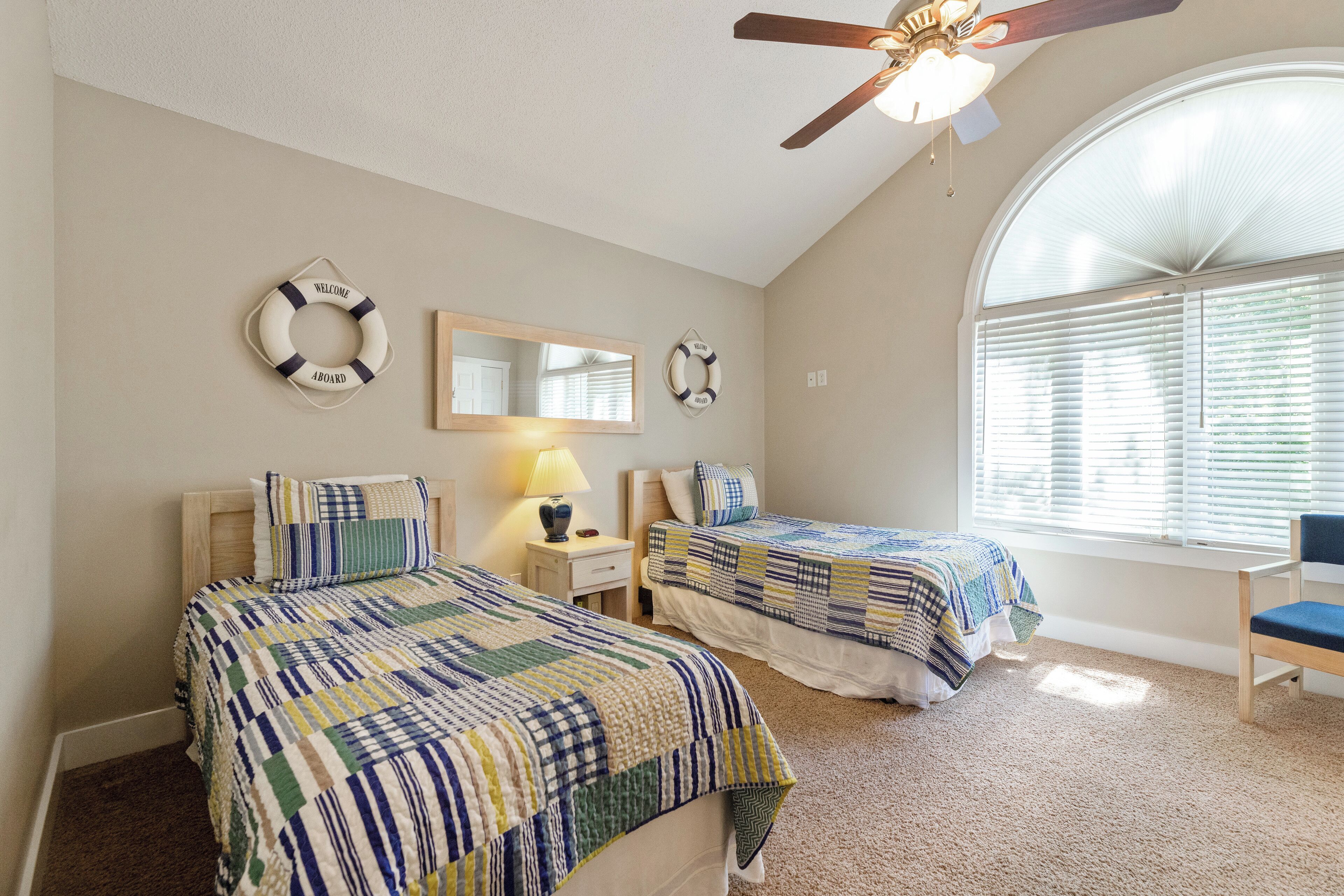 A bedroom with two beds, a ceiling fan, a large window with blinds, and a wall-mounted mirror.