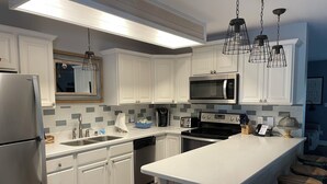 A modern kitchen with white cabinets, a stainless steel refrigerator, a microwave, a stove, and a sink. There are bar stools and a trash bin. - Four Bedroom Yacht Condo (Grand Rivers)