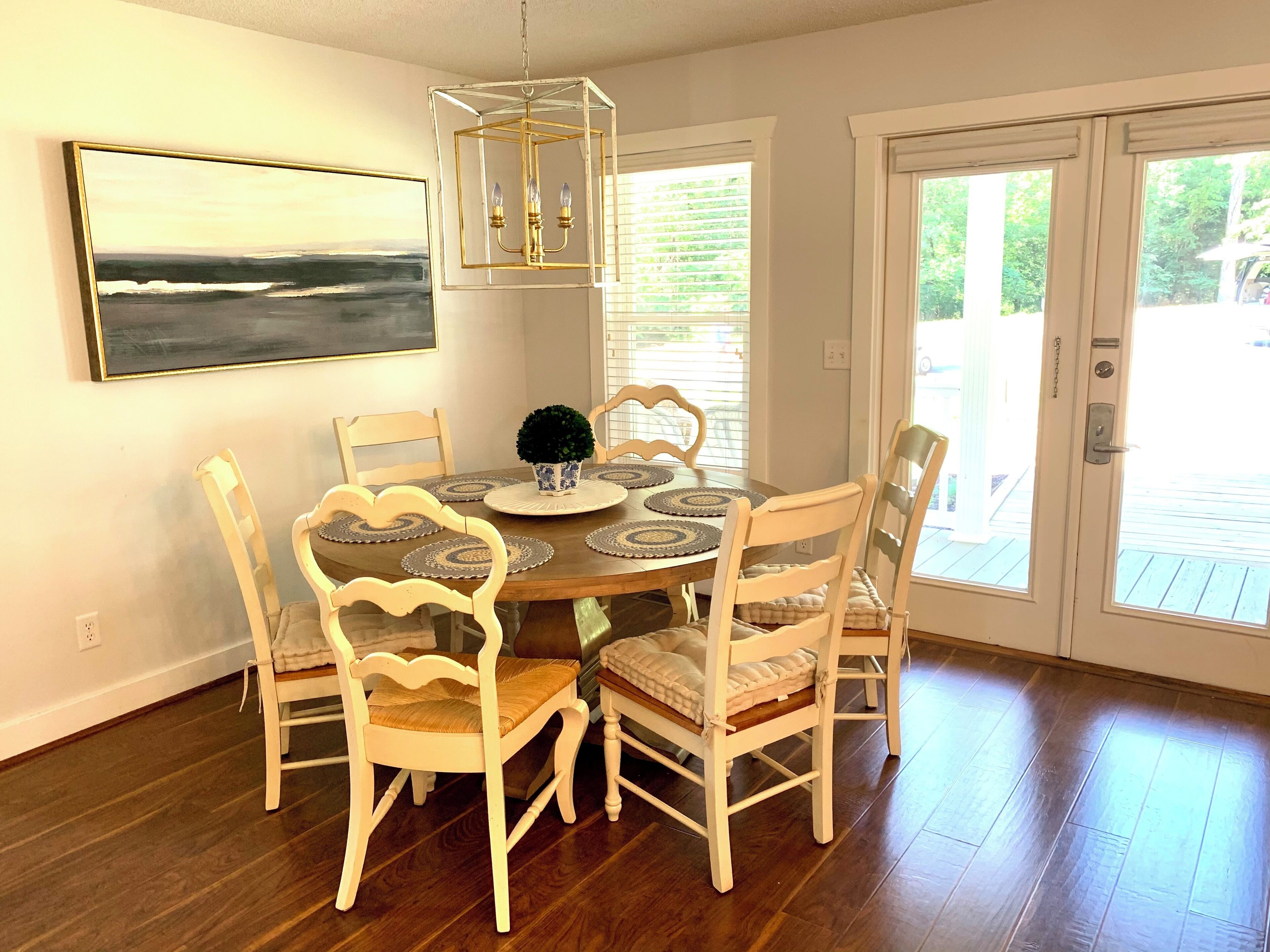 A dining area with a wooden table, six chairs, a chandelier, a potted plant, and a painting on the wall.