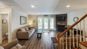 A living room with a sofa, coffee table, and a fireplace. There is a staircase with a wooden railing and a door labeled "Lake House". - Four Bedroom Yacht Condo (Grand Rivers)