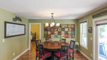 A dining area with a round wooden table surrounded by six chairs with red cushions, a chandelier, and a wall decorated with various framed artworks.