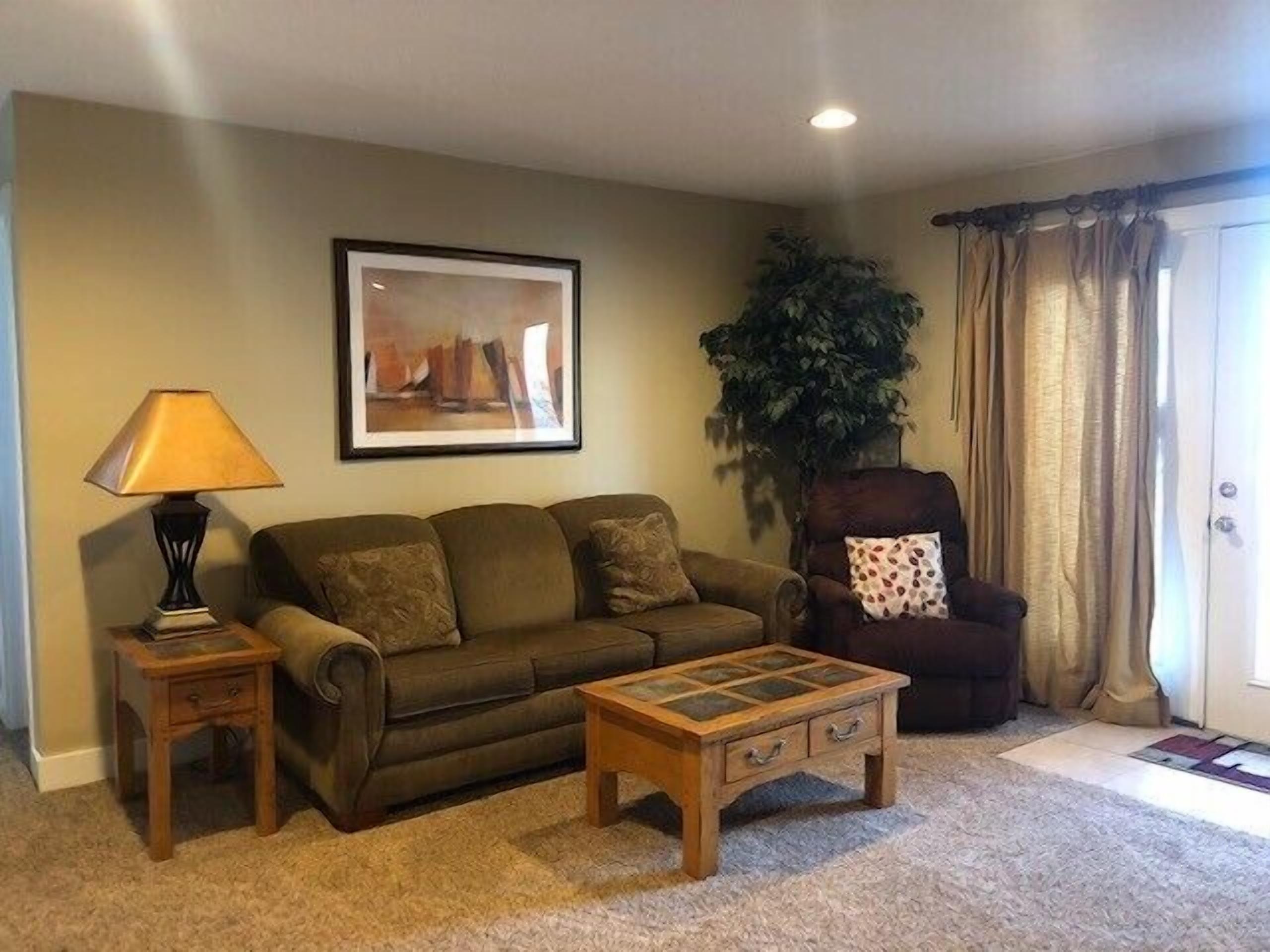 A living room with a sofa, a coffee table, a lamp, a chair, a plant, and a door.
