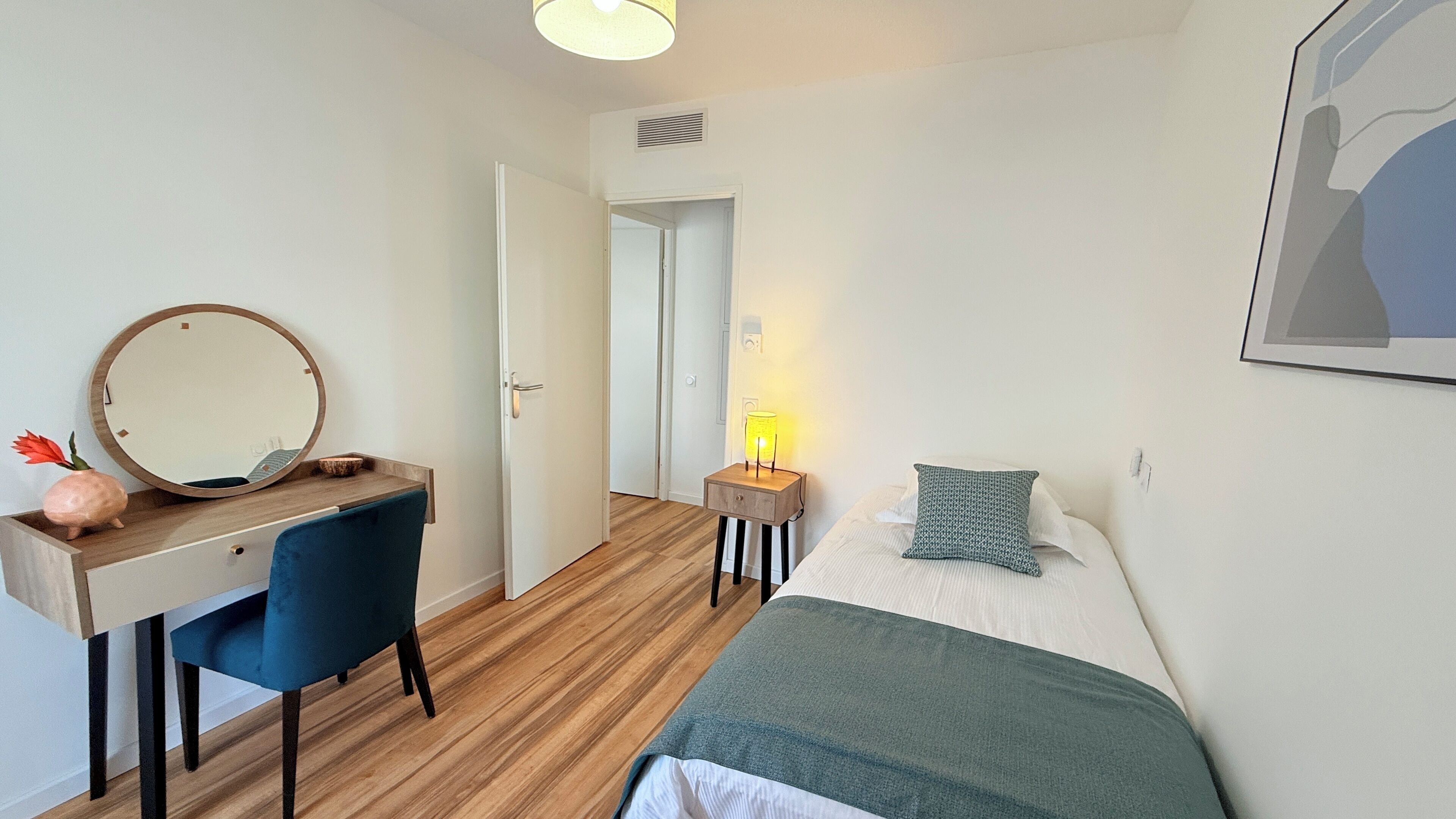 Une chambre à coucher avec un lit, un bureau doté d’un miroir, une table de chevet avec une lampe et une porte donnant sur une autre pièce.