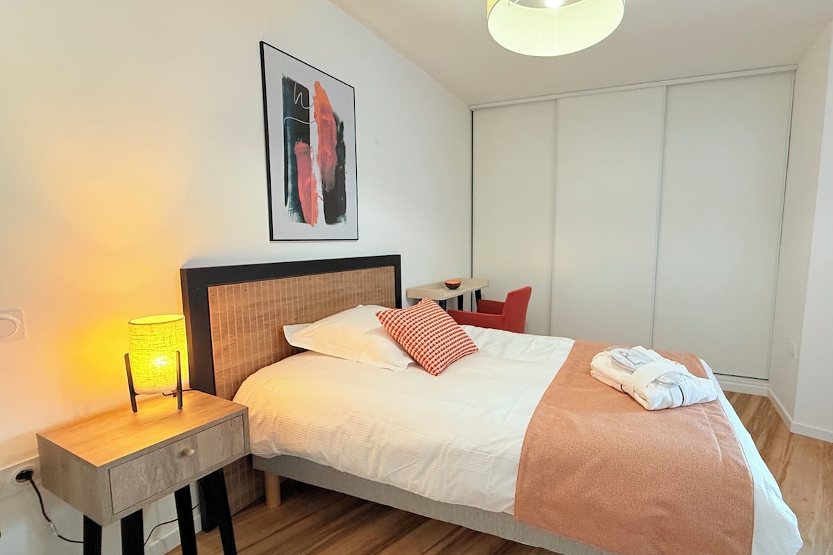 A modern hotel room with a wooden bed, a bedside table with a lamp, a framed artwork on the wall, and a window with curtains.
