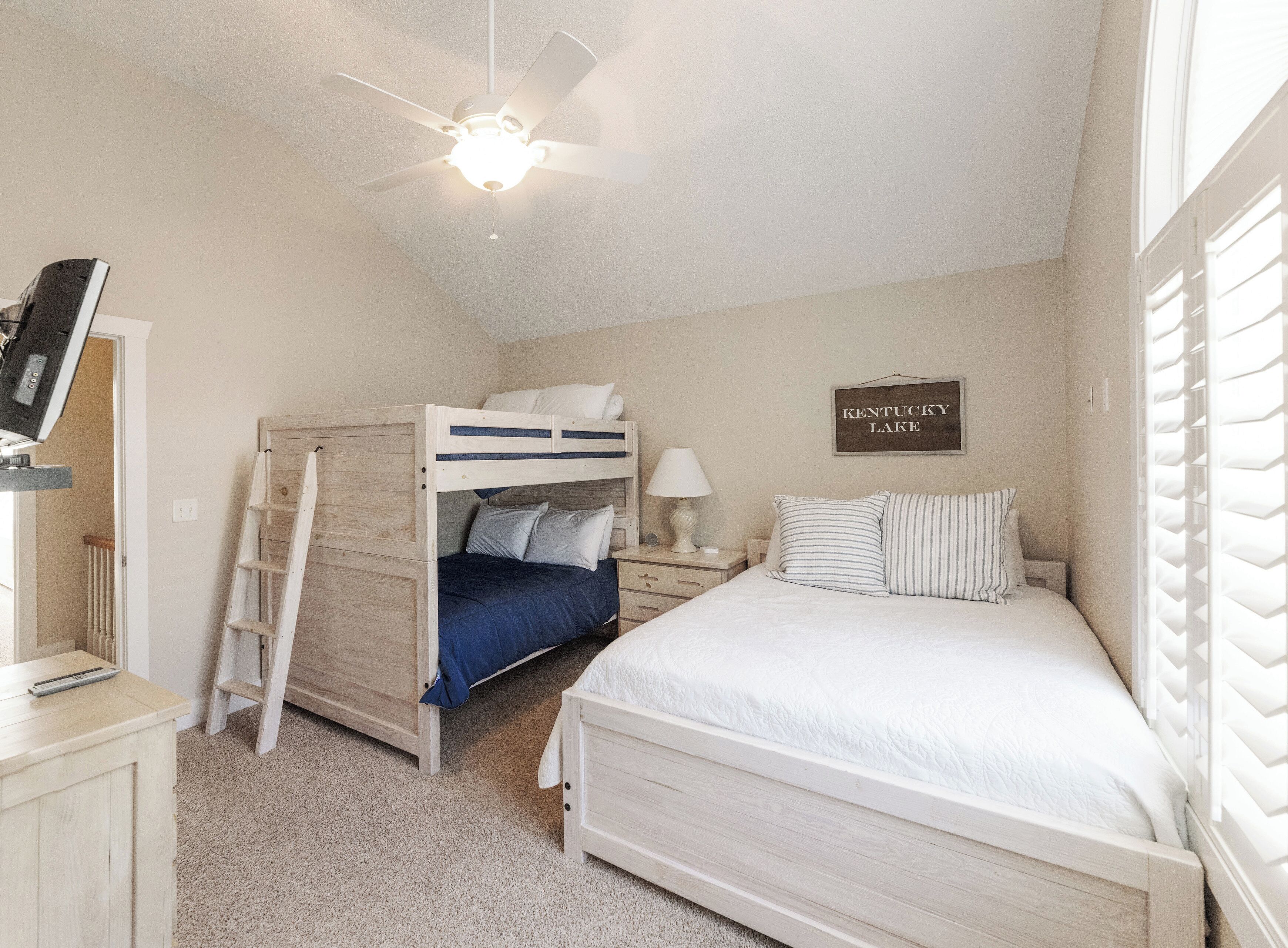 A bedroom with a bunk bed, a single bed, a ceiling fan, a bedside table with a lamp, a sign on the wall, and a TV on a stand.