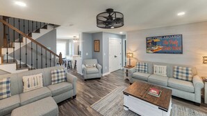 A spacious living room with a staircase, a ceiling fan, a couch with plaid cushions, a coffee table, and a wall-mounted sign. - Four Bedroom Yacht Condo (Grand Rivers)